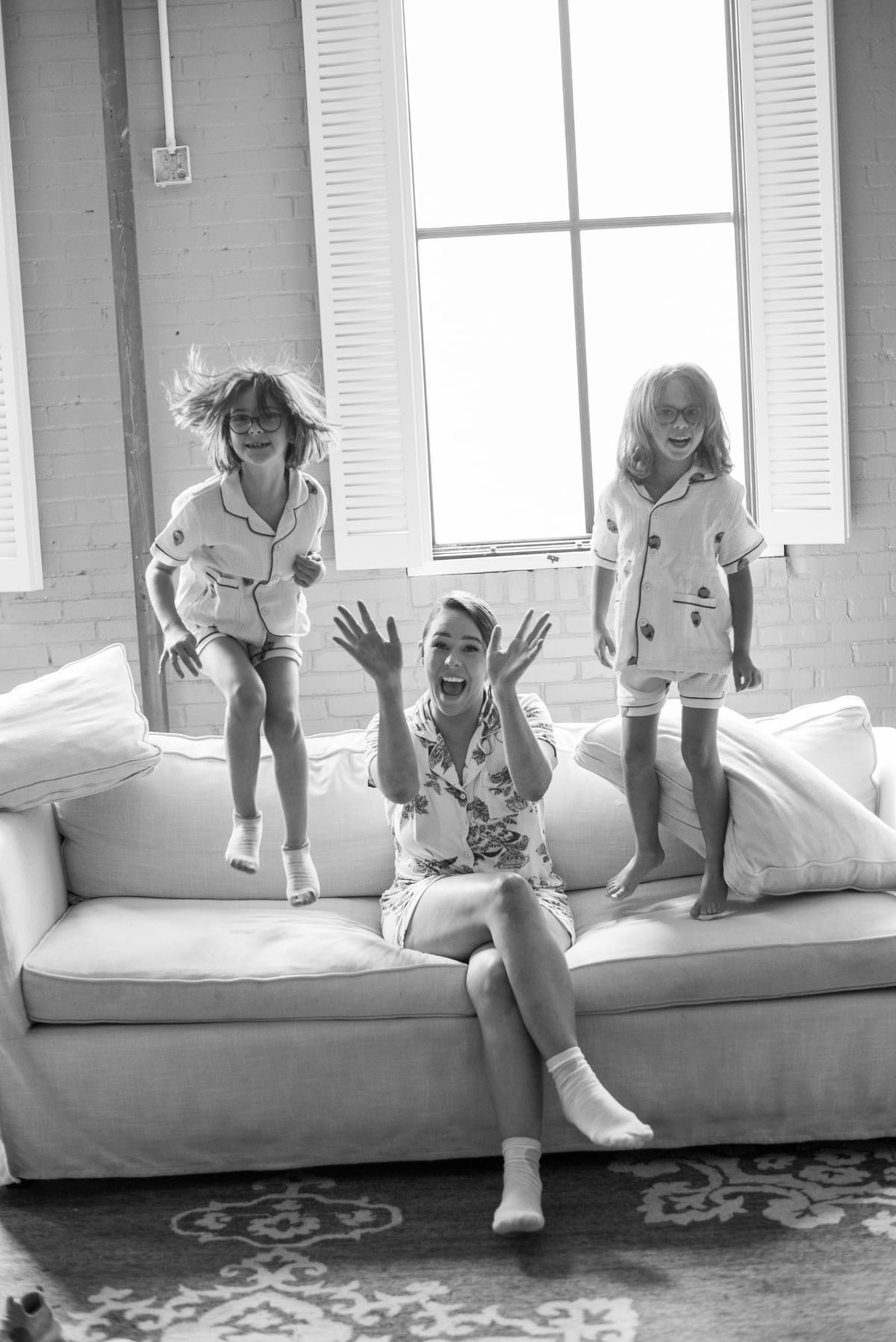 Préparatifs joyeux dans un loft : deux fillettes en pyjama sautent sur le canapé autour de la mariée, en noir et blanc