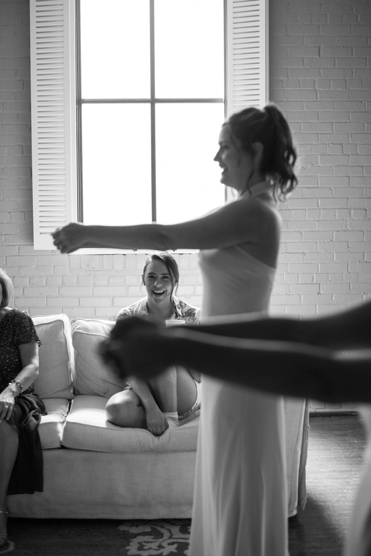 Mariée debout au milieu de ses amies pendant les préparatifs, grande fenêtre en fond, en noir et blanc