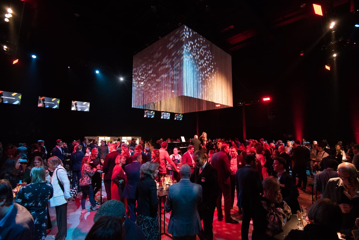 Grande salle plongée dans le rouge, cube lumineux suspendu au-dessus de la foule d'un cocktail